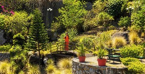 Un jardín con muro de piedra, plantas en macetas, un puente de madera y mobiliario de exterior.