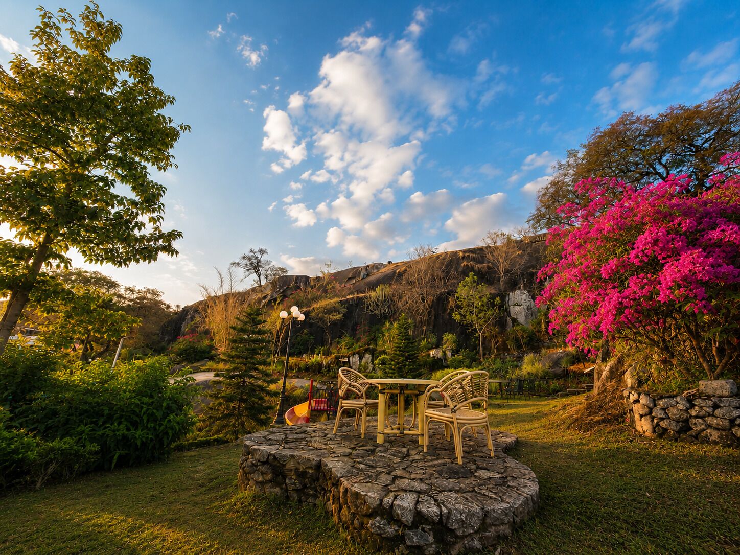 Un jardin avec une table et des chaises en pierre, entouré d’une végétation luxuriante et d’un arbre à fleurs roses.