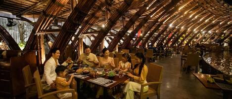 Um restaurante com estrutura de telhado em bambu peculiar e área de refeições com cadeiras de vime e uma mesa comprida.