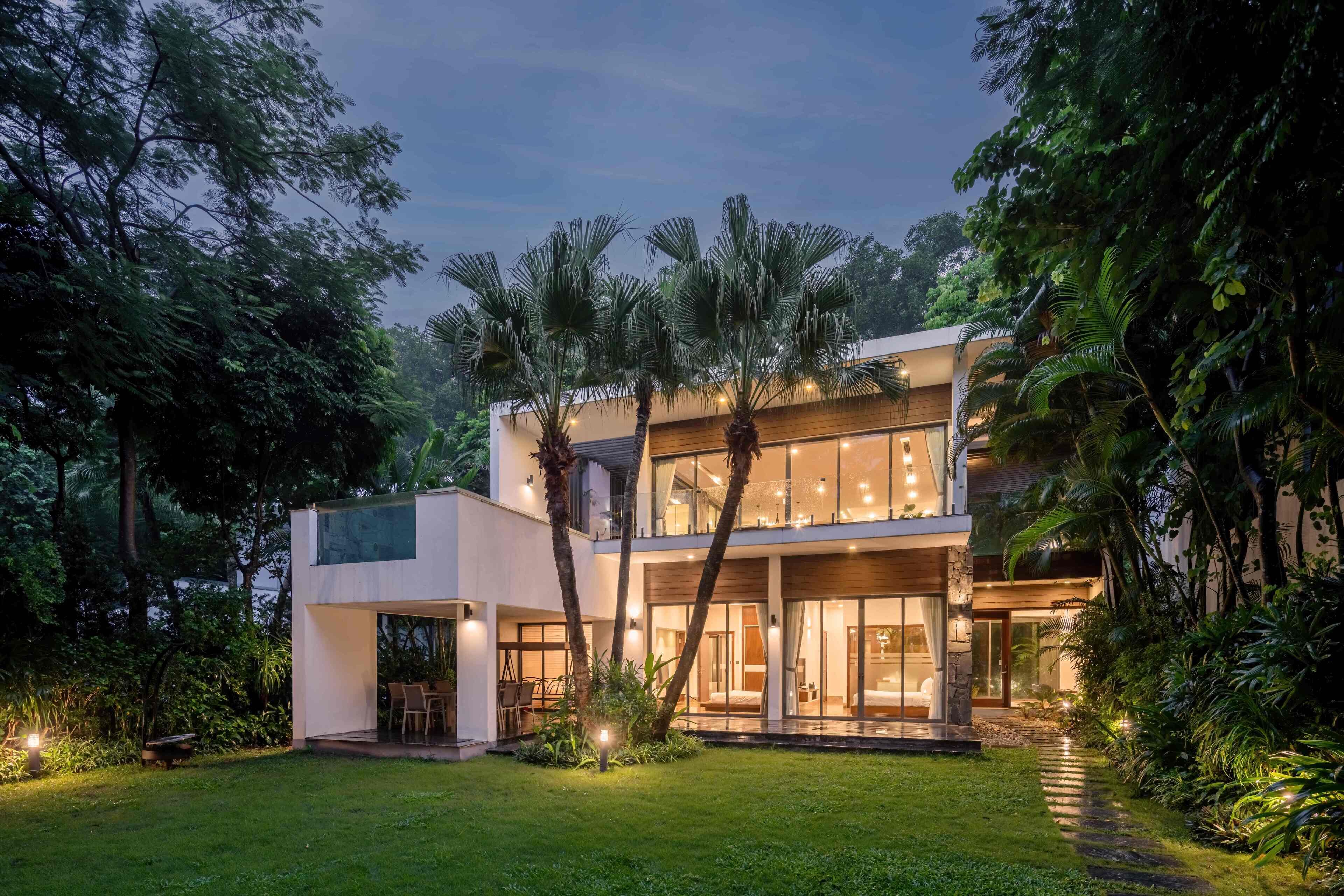 A modern two-story house with a glass facade, surrounded by lush greenery and palm trees.