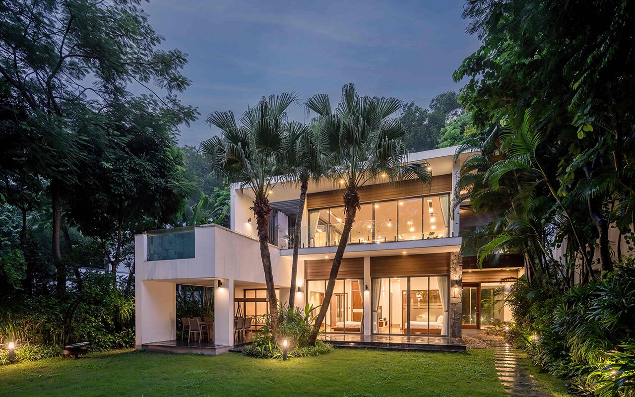 Una casa moderna de dos pisos con fachada de vidrio, rodeada de exuberante vegetación y palmeras.