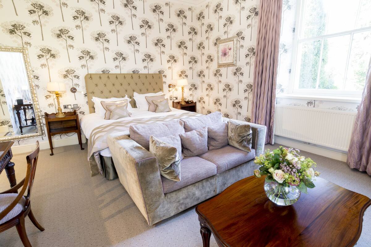 A bedroom with a large bed, a sofa, a wooden nightstand, a desk, and a window with curtains.
