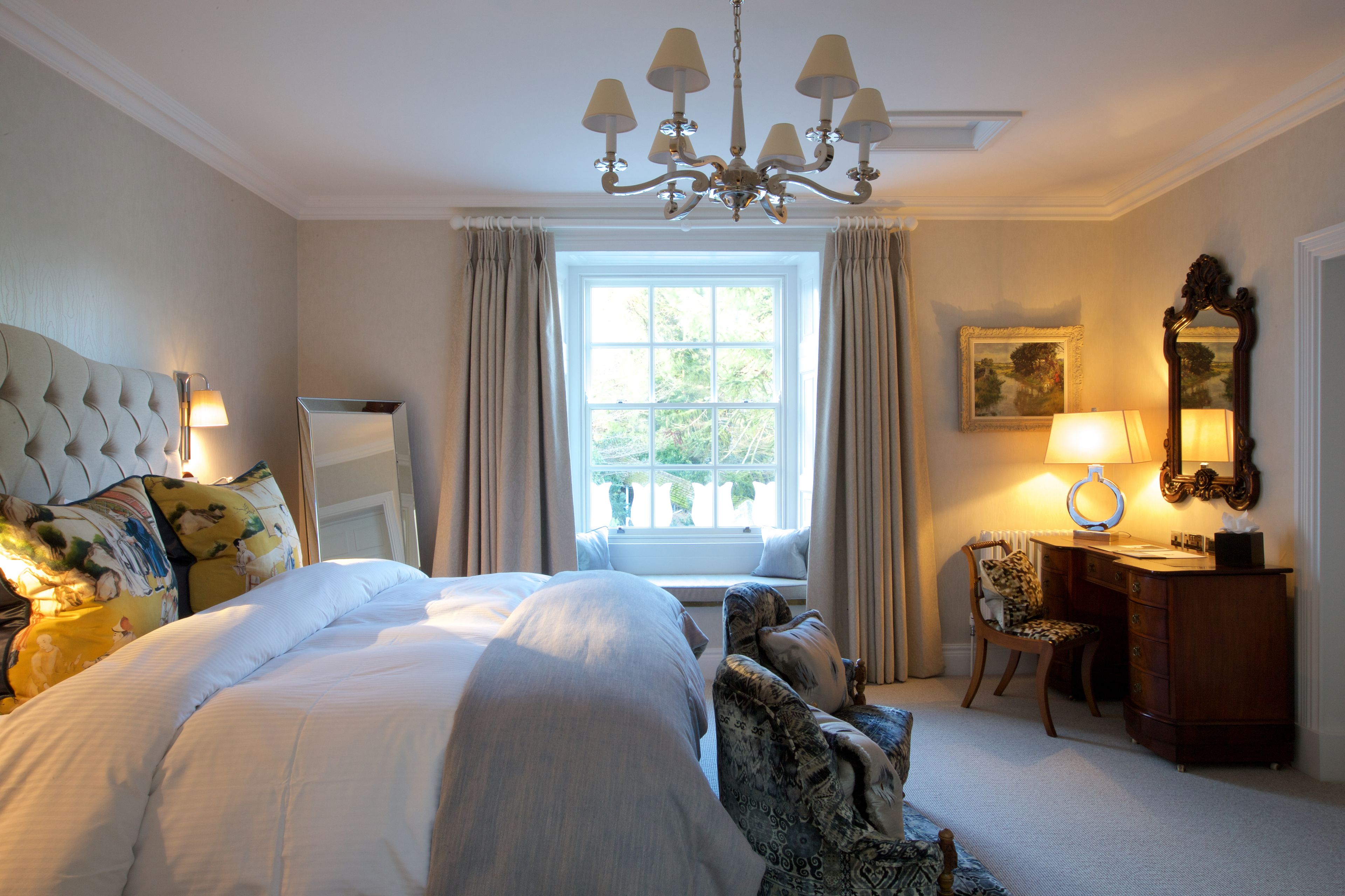 A well-lit bedroom with a large bed, a desk with a chair, a chandelier, and a window with curtains.