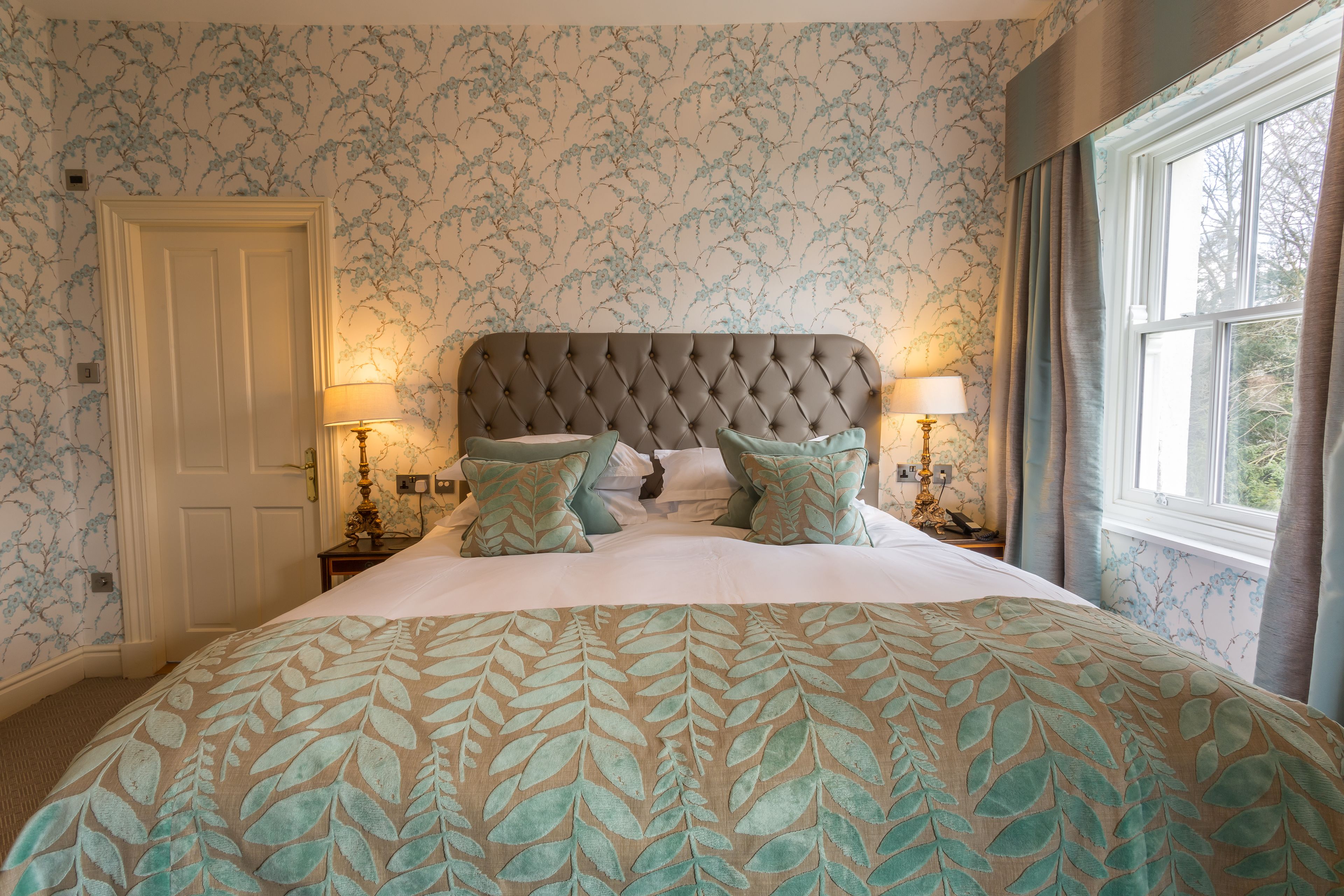 A bedroom with a tufted headboard, floral wallpaper, and a bed with a leaf-patterned bedspread. There are two bedside lamps and a window with curtains.