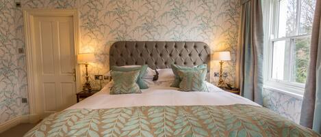 A bedroom with a tufted headboard, floral wallpaper, and a bed with a leaf-patterned bedspread. There are two bedside lamps and a window with curtains.