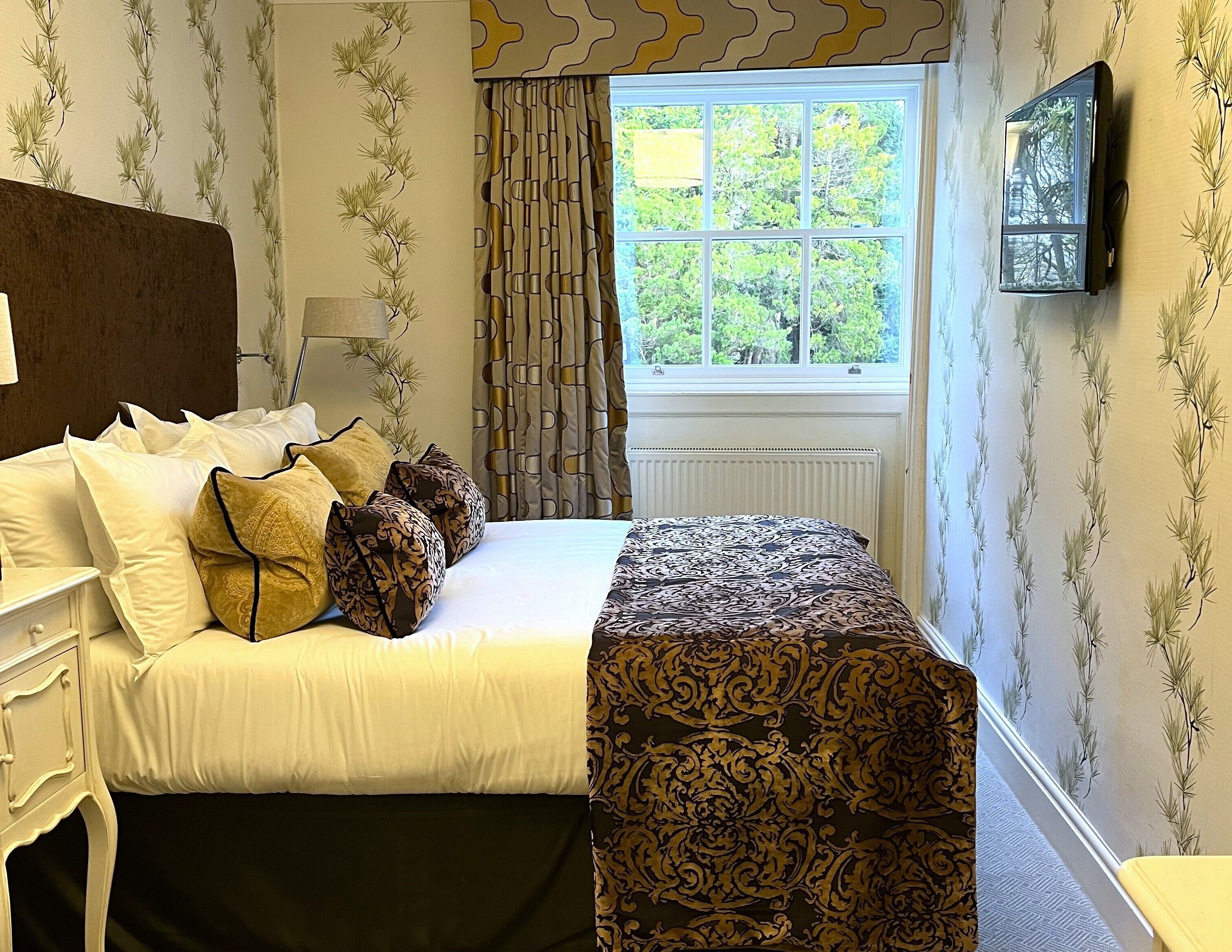 A neatly made bed with white and patterned bedding, a bedside table with a lamp, and a window with curtains.
