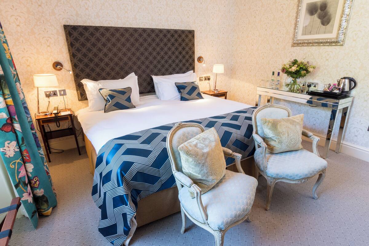 A hotel room with a large bed, two bedside tables, a nightstand, two upholstered chairs, a small desk with a mirror, and a wall-mounted picture.