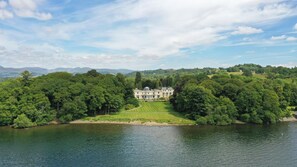 Un vaste manoir historique, entouré d’une végétation luxuriante et bordé d’un lac.