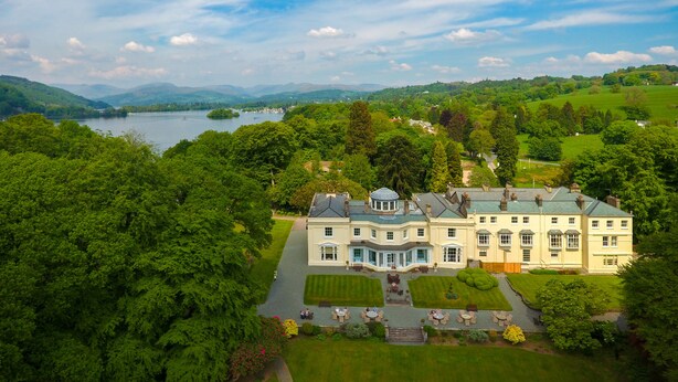 A large, historic building with a symmetrical facade, multiple windows, and a central dome. It is surrounded by lush greenery and a lake, with hills in the background. - Storrs Hall Hotel (Windermere)