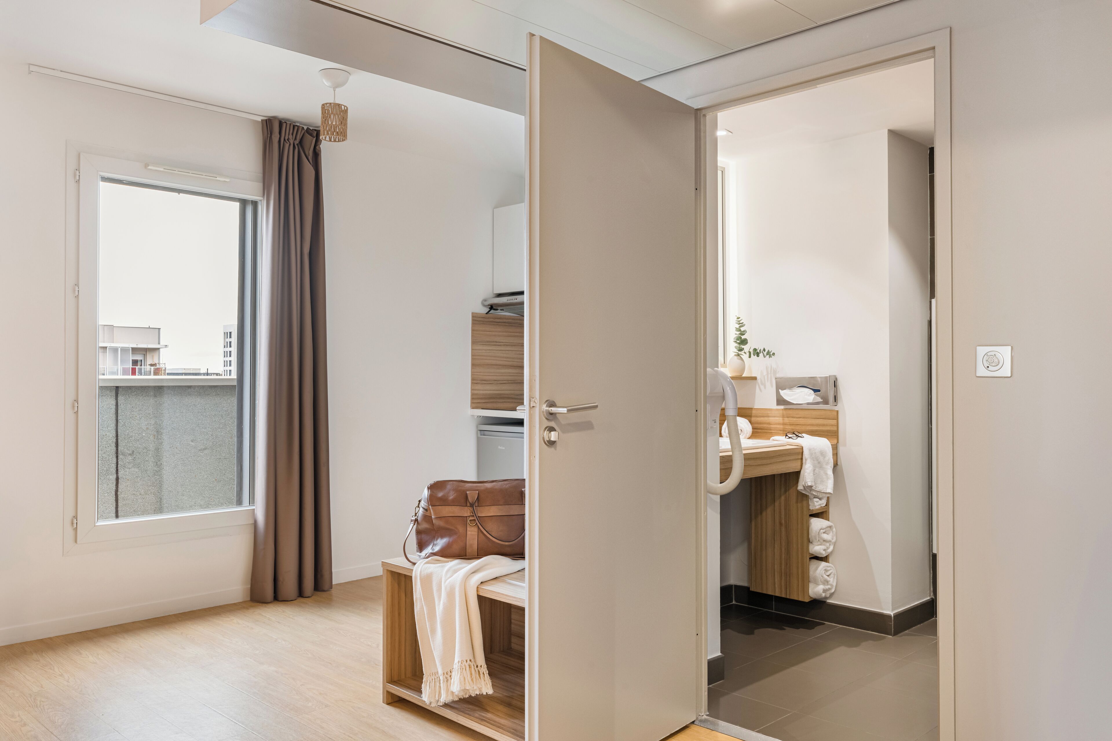 Eine moderne Wohnung mit großem Fenster, einer Holzbank, einer braunen Ledertasche und einer Küchenzeile mit Spüle und Handtuchhalter.