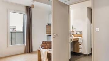 Eine moderne Wohnung mit großem Fenster, einer Holzbank, einer braunen Ledertasche und einer Küchenzeile mit Spüle und Handtuchhalter.