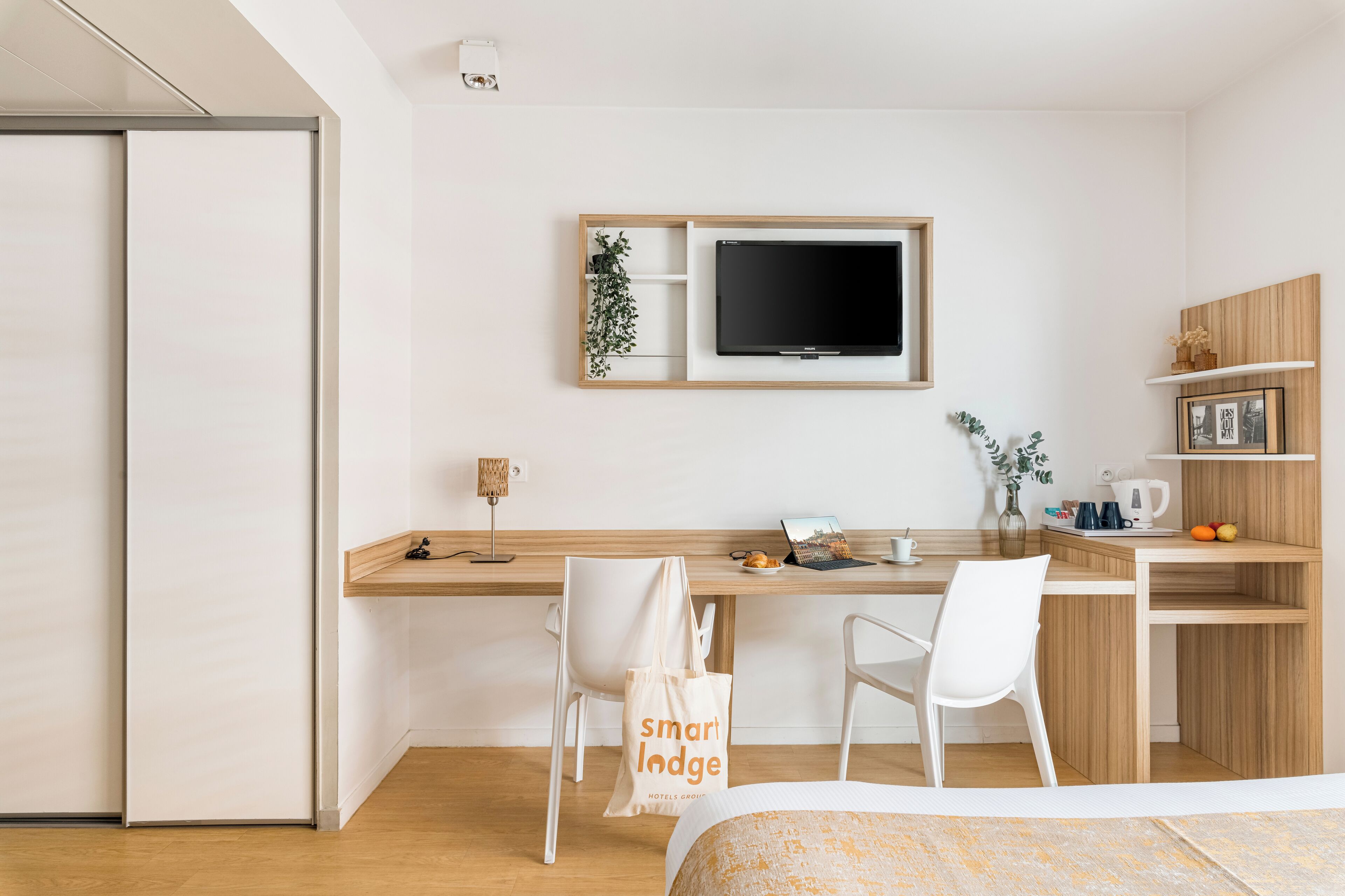 Une chambre d’hôtel moderne équipée d’un lit, d’un bureau avec un ordinateur, d’une télévision et d’une petite kitchenette.