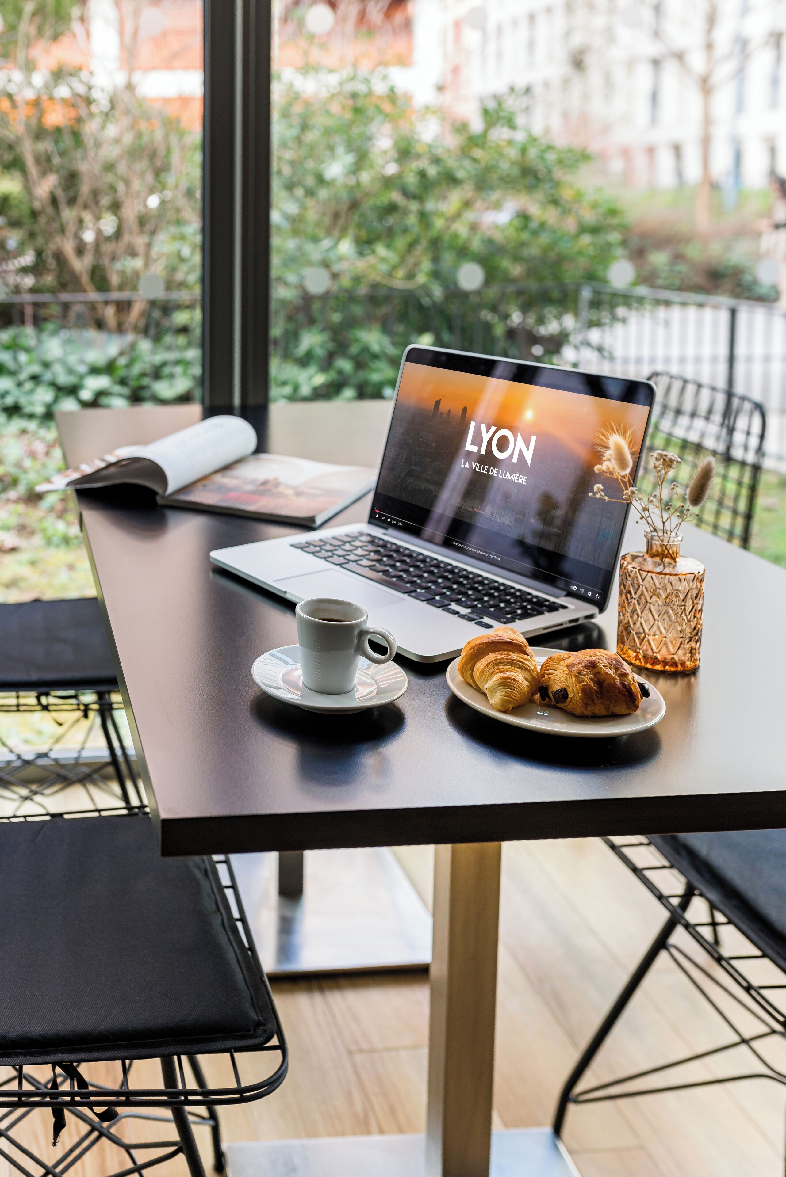 Een laptop op een tafel met een kop koffie, een bord croissants en een decoratieve vaas.