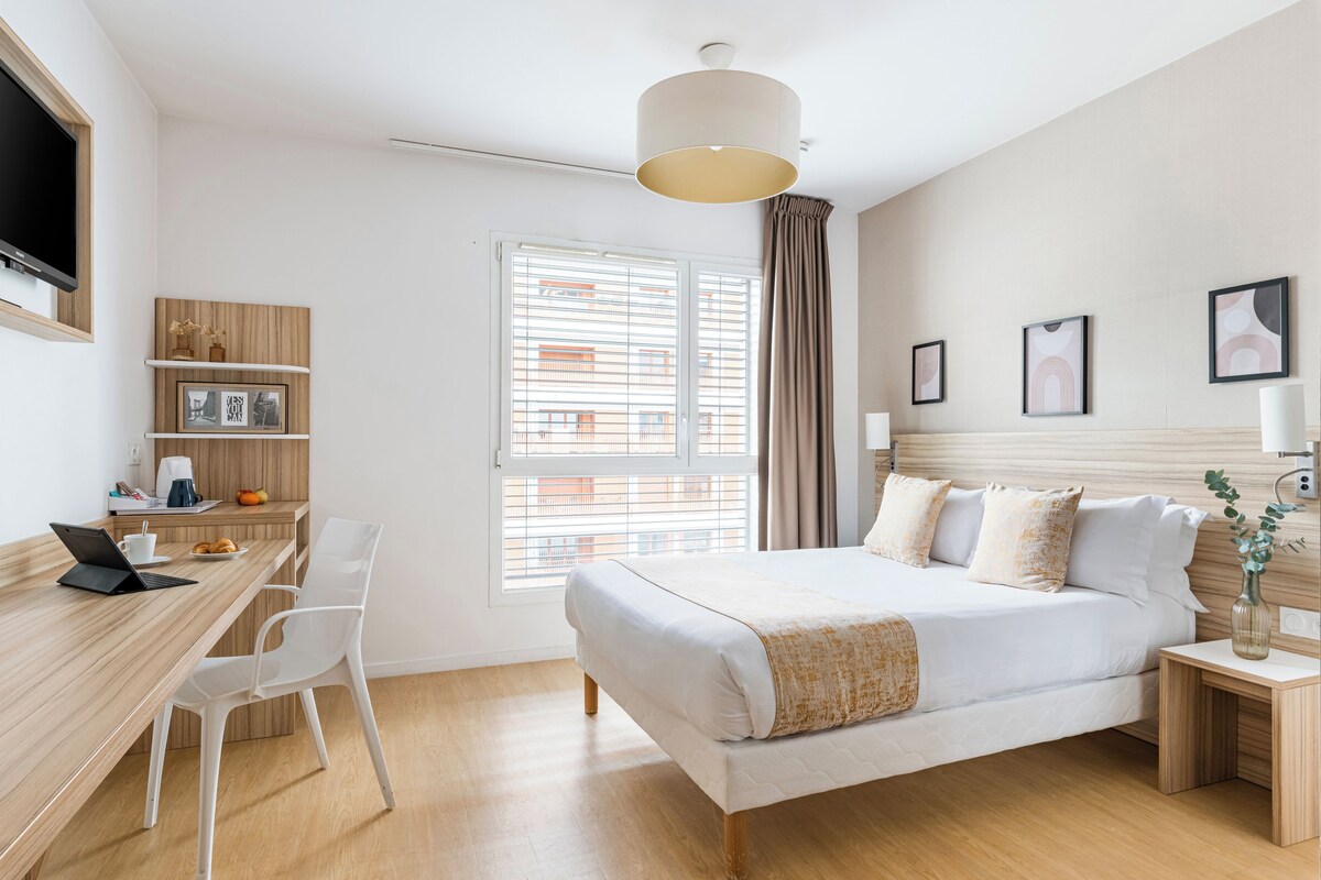 A modern hotel room with a bed, a desk with a laptop, a chair, a window with blinds, and a TV mounted on the wall.