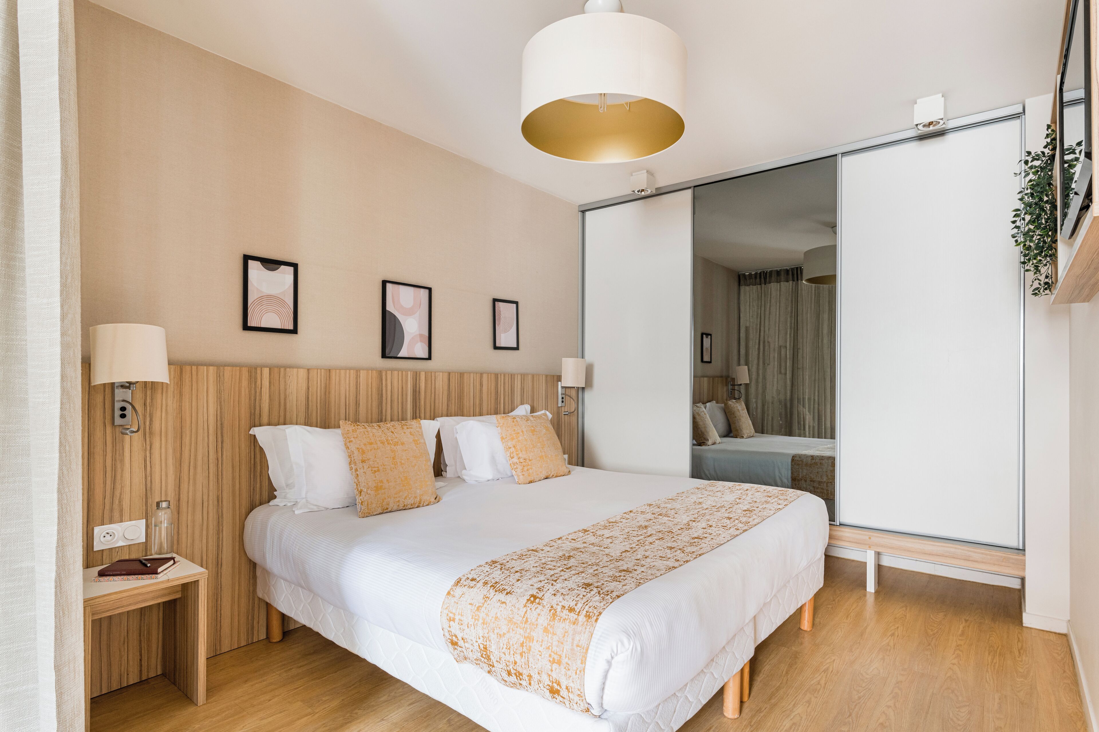 A modern hotel room with a large bed, bedside lamps, a wooden headboard, a large mirror, and a sliding door closet.