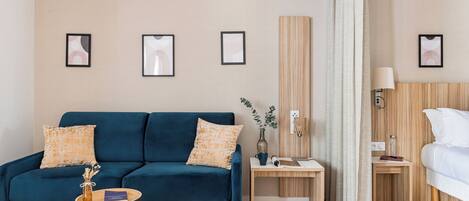 Una habitación de hotel moderna con un sofá azul, una mesita de noche de madera, una cama y cuadros en las paredes.