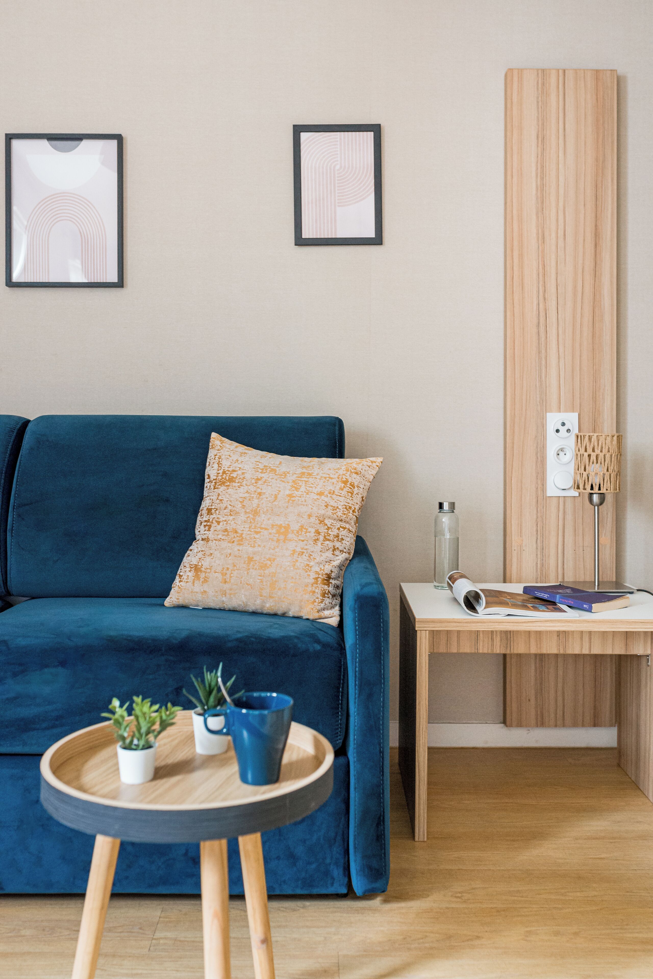 A modern living room with a blue sofa, a small wooden side table, two framed pictures on the wall, and a wooden panel.