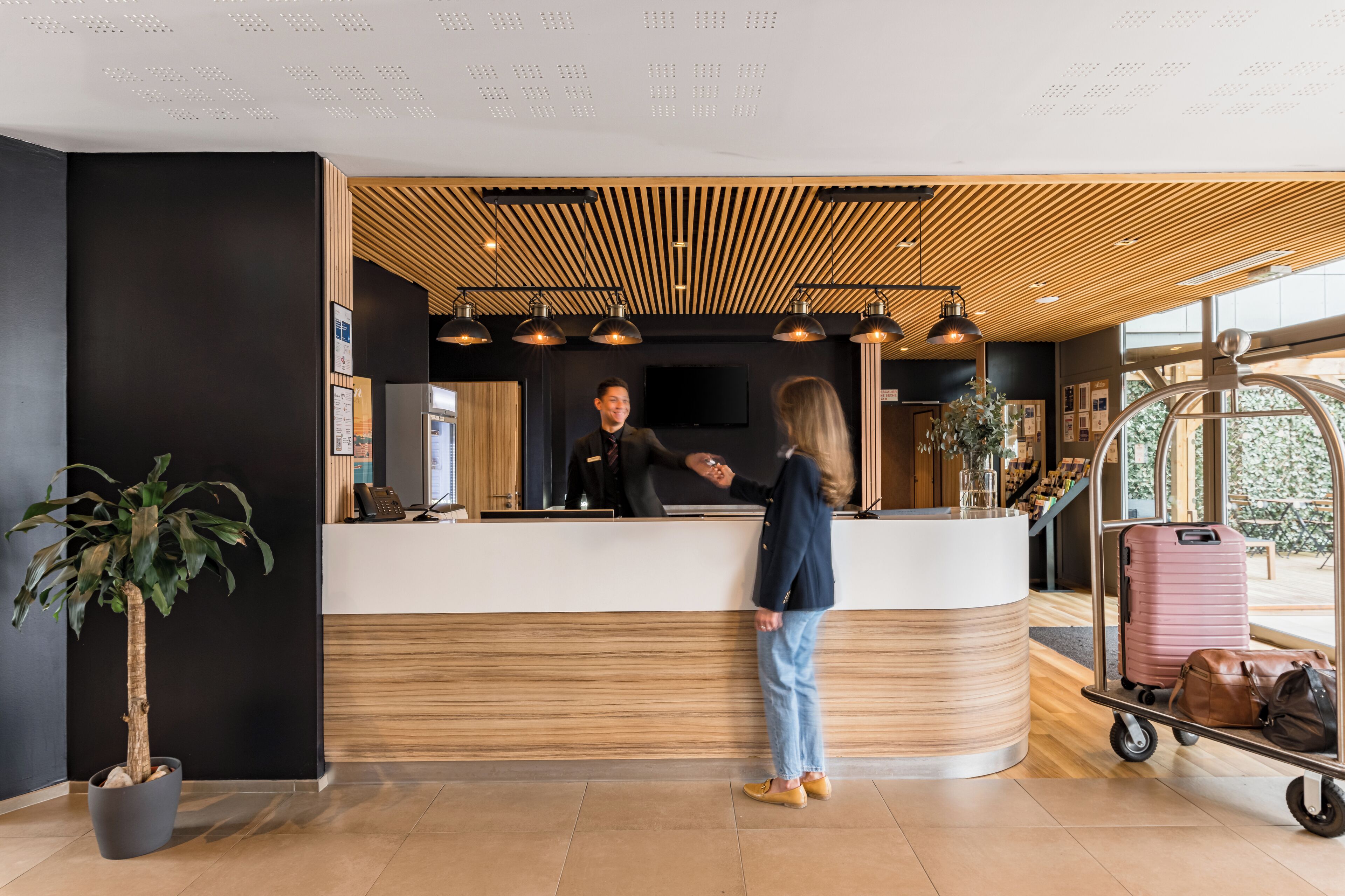 A reception desk with a modern design, a potted plant, and luggage on a cart.