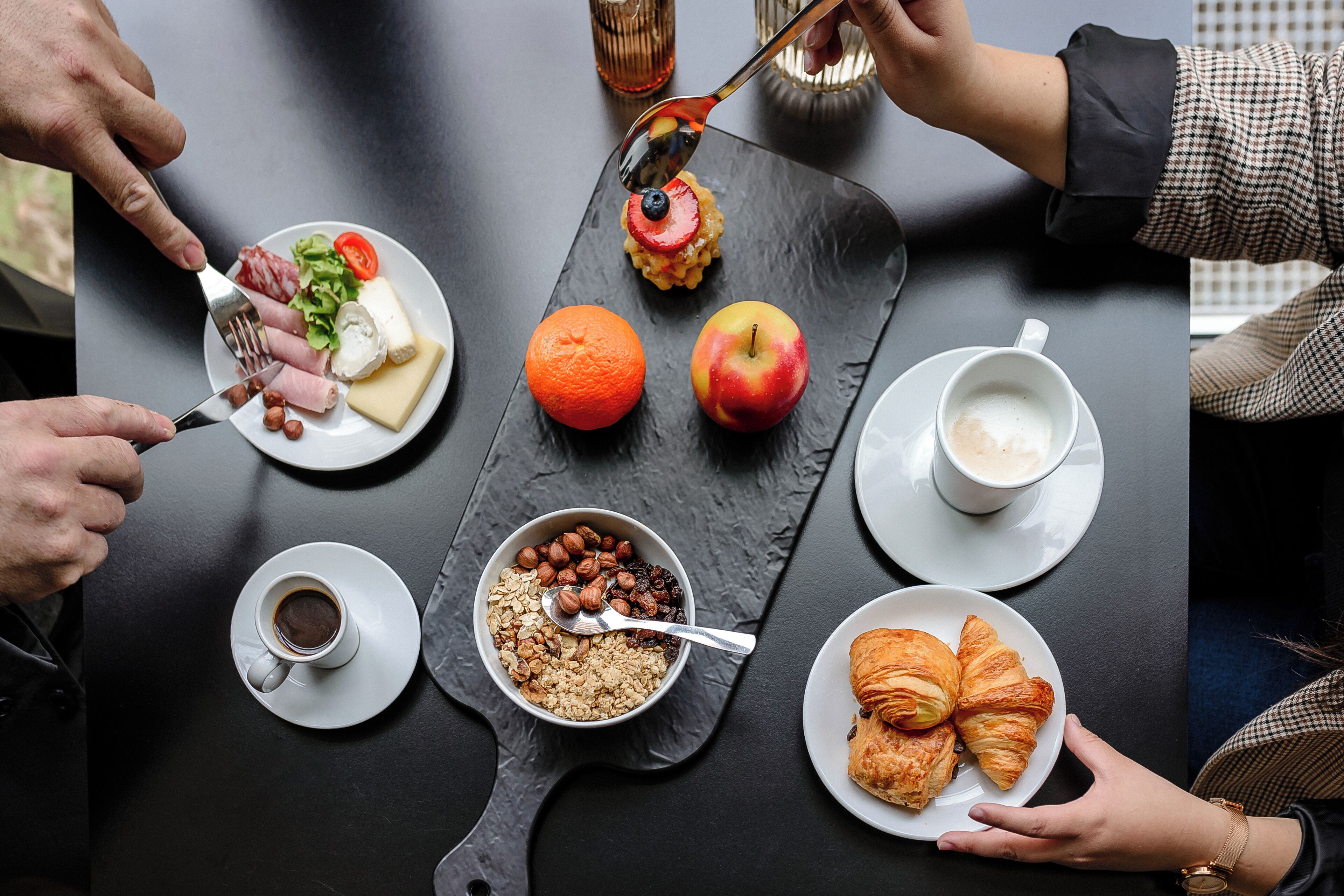 Una mesa de comedor con diversos alimentos y bebidas, incluyendo un plato con queso, frutos secos y un tazón de cereal, un plato con croissants y una taza de café.