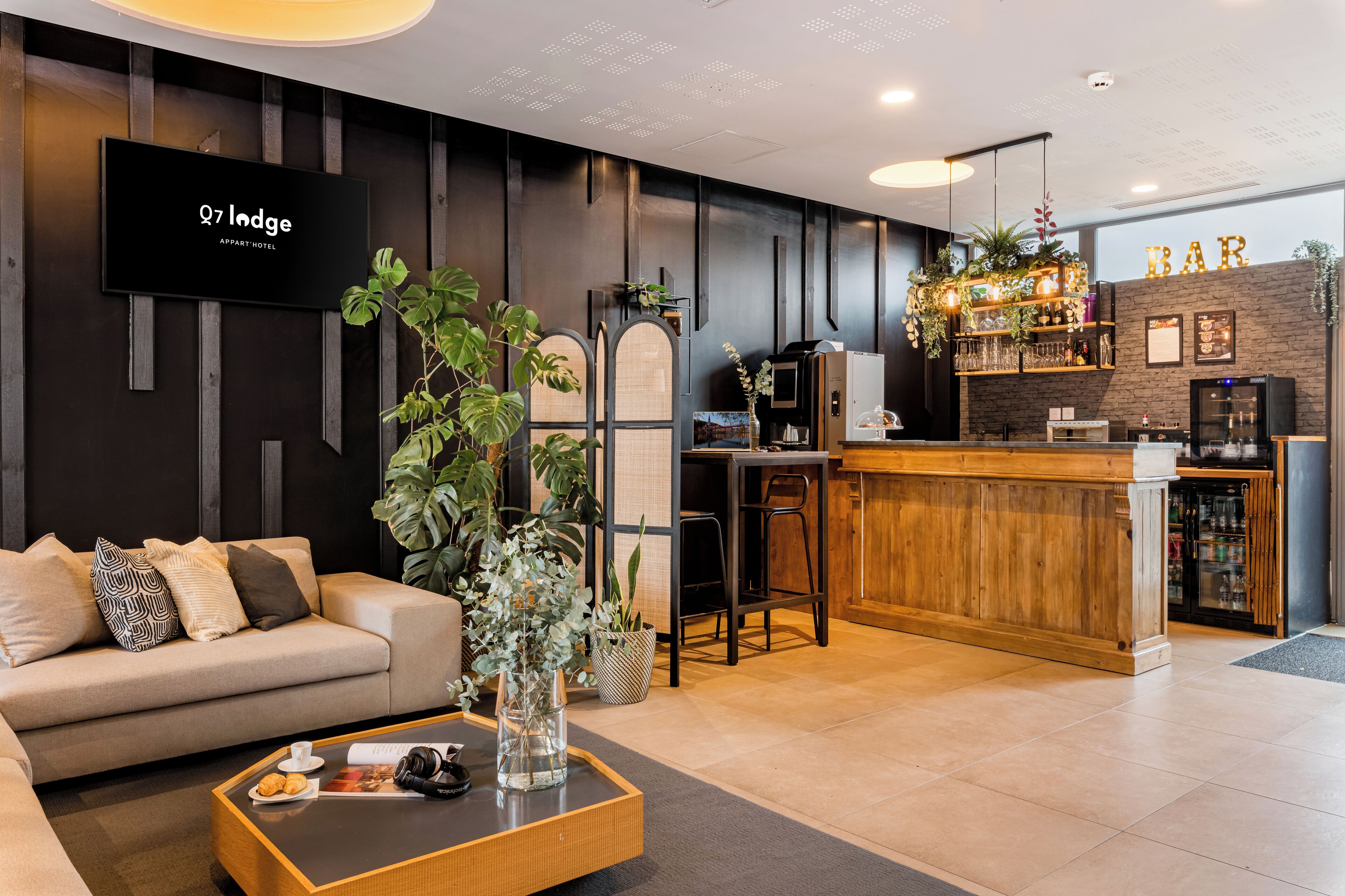 A modern hotel lobby with a sofa, coffee table, TV, bar area, and a reception desk.