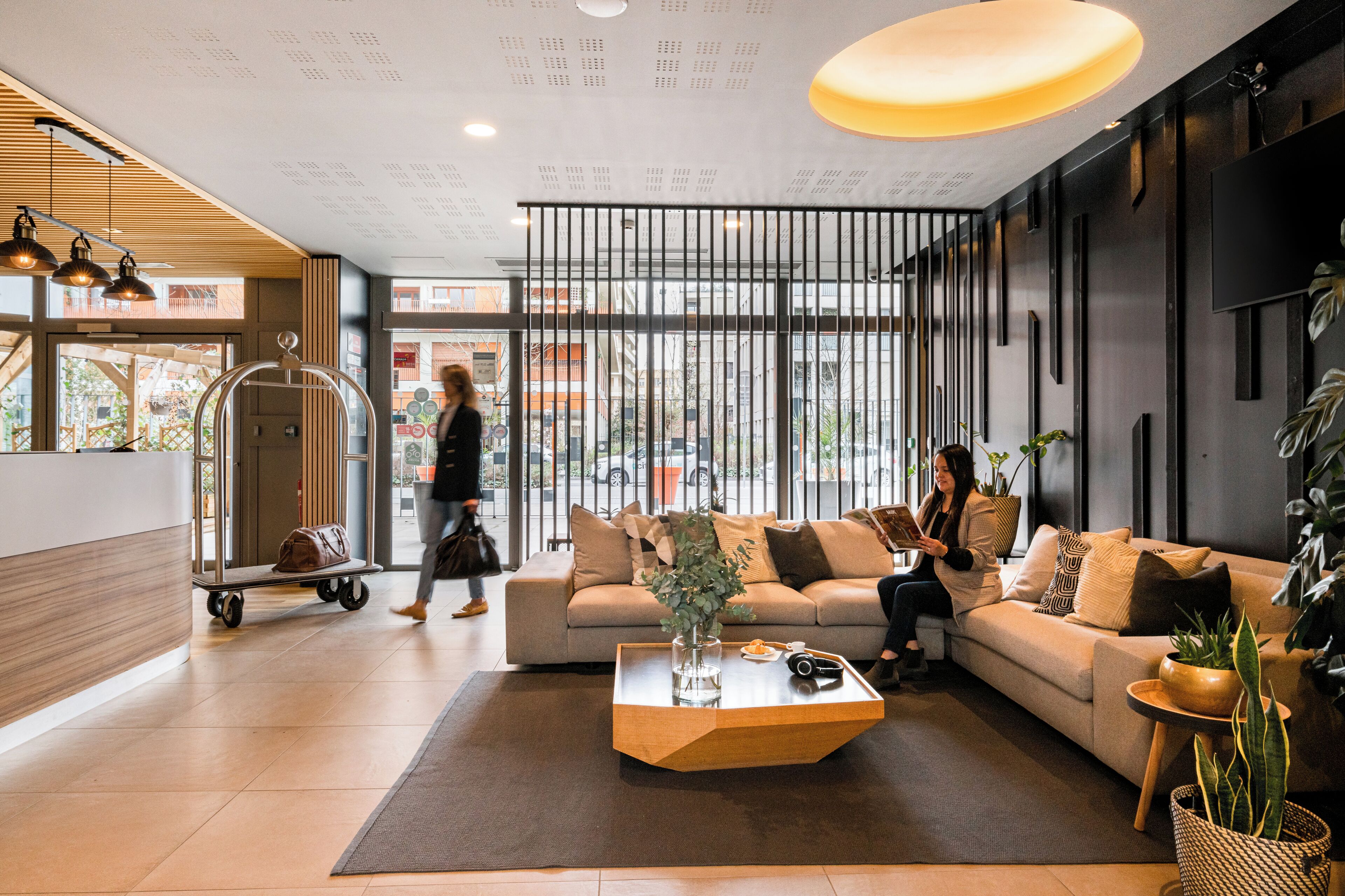 A modern lobby with a reception desk, a seating area with a sofa and armchairs, a coffee table, and potted plants.