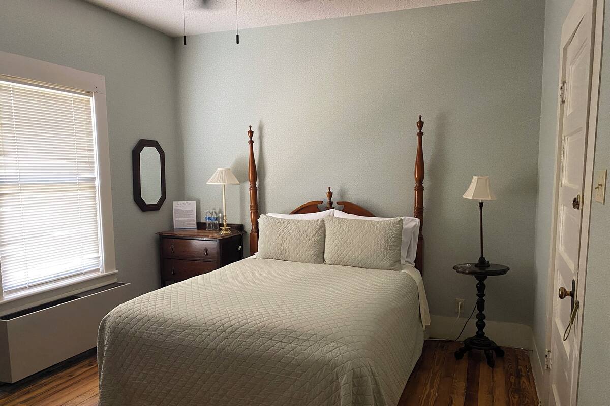A bedroom with a four-poster bed, a nightstand, a bedside table, a lamp, and a window with blinds.