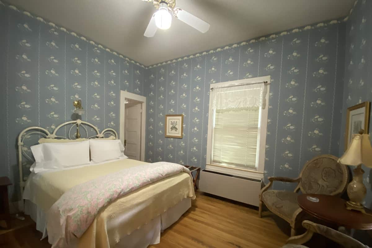 A bedroom with a bed, a chair, a window with blinds, a ceiling fan, and a radiator.