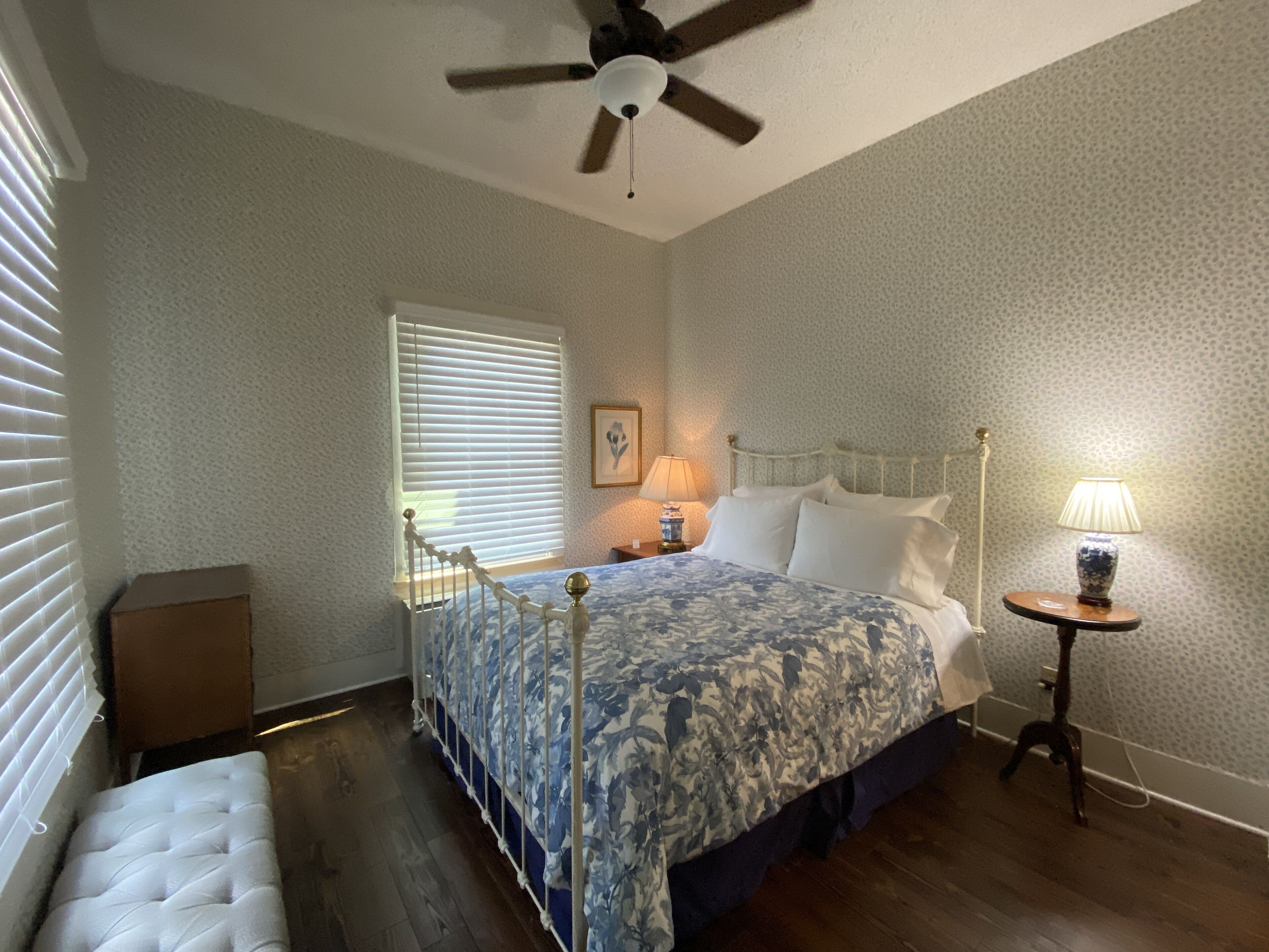A bedroom with a bed, a ceiling fan, a bedside lamp, a nightstand, a window with blinds, and a small table with a lamp.