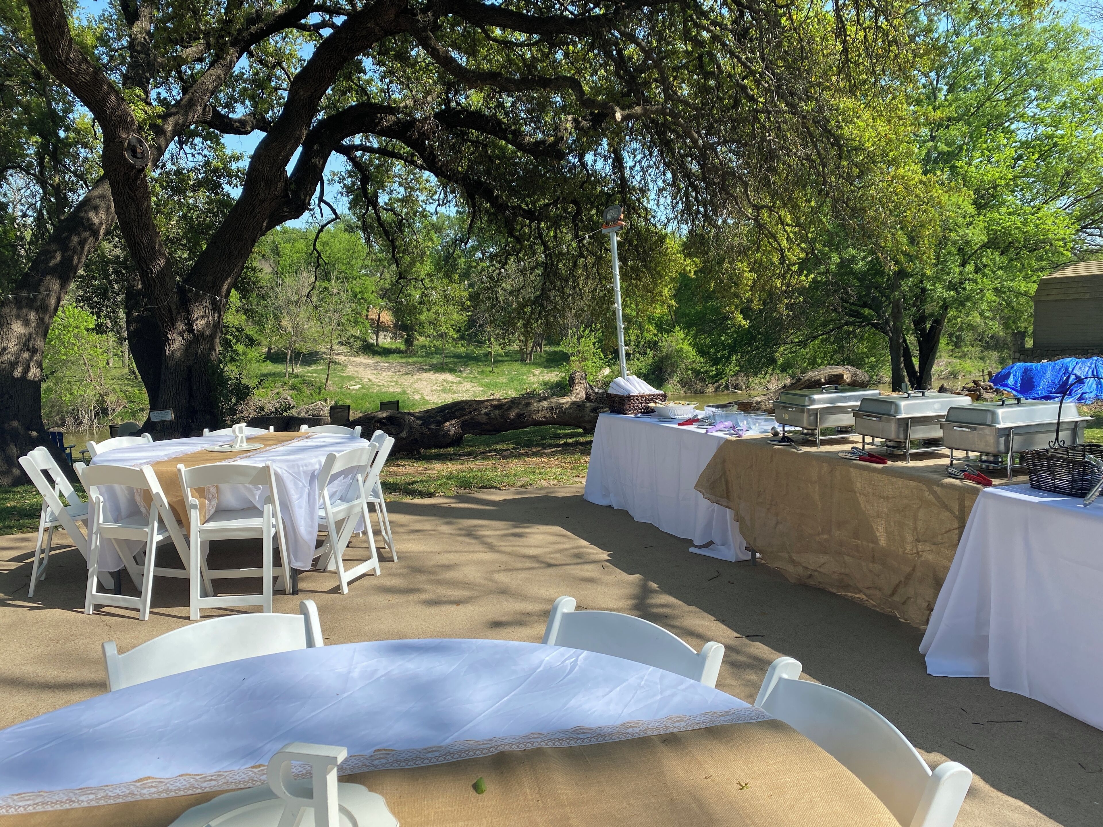 An outdoor dining area with white tables and chairs, surrounded by trees and a natural landscape.