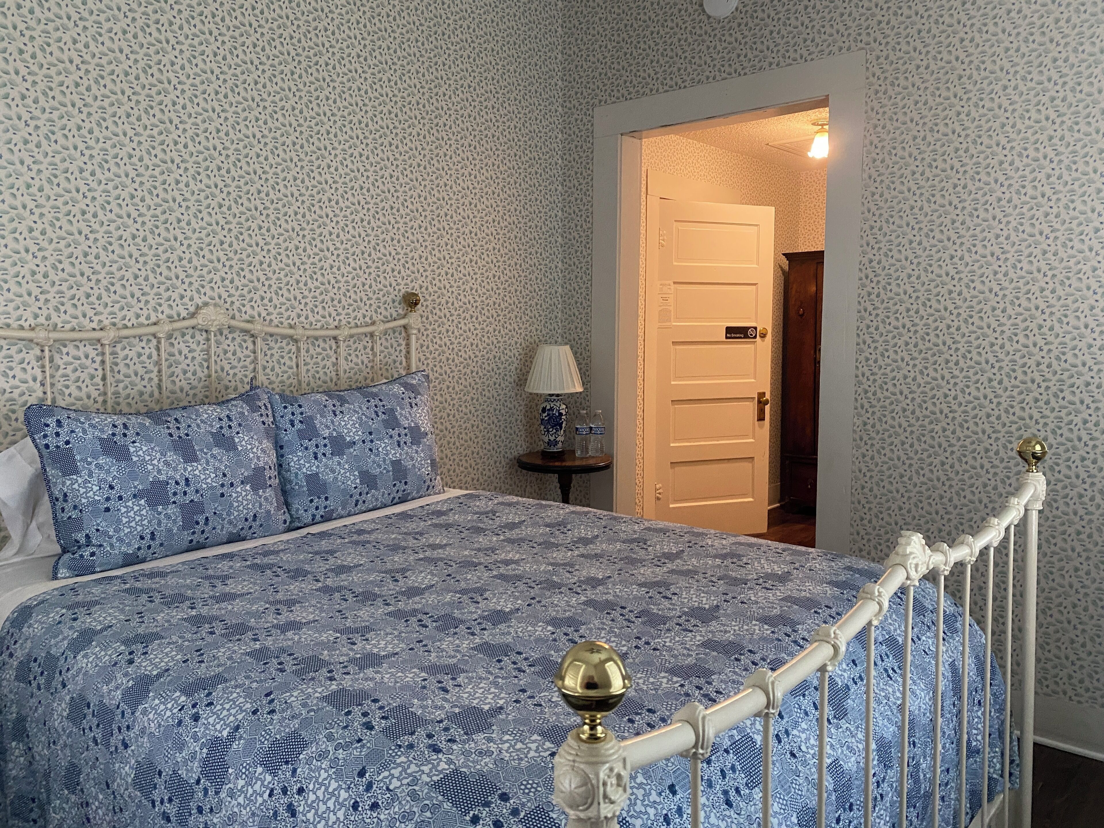 Un dormitorio con una colcha estampada, una cama con estructura de metal, una mesita de noche con una lámpara y una puerta que da a otra habitación.