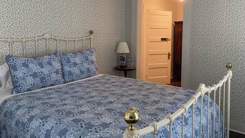 Un dormitorio con una colcha estampada, una cama con estructura de metal, una mesita de noche con una lámpara y una puerta que da a otra habitación.