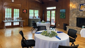 A spacious room with wooden walls, a stone fireplace, and a ceiling fan. There are tables set for dining and a kitchen area with a sink and countertop. - Inn On The River (Glen Rose)