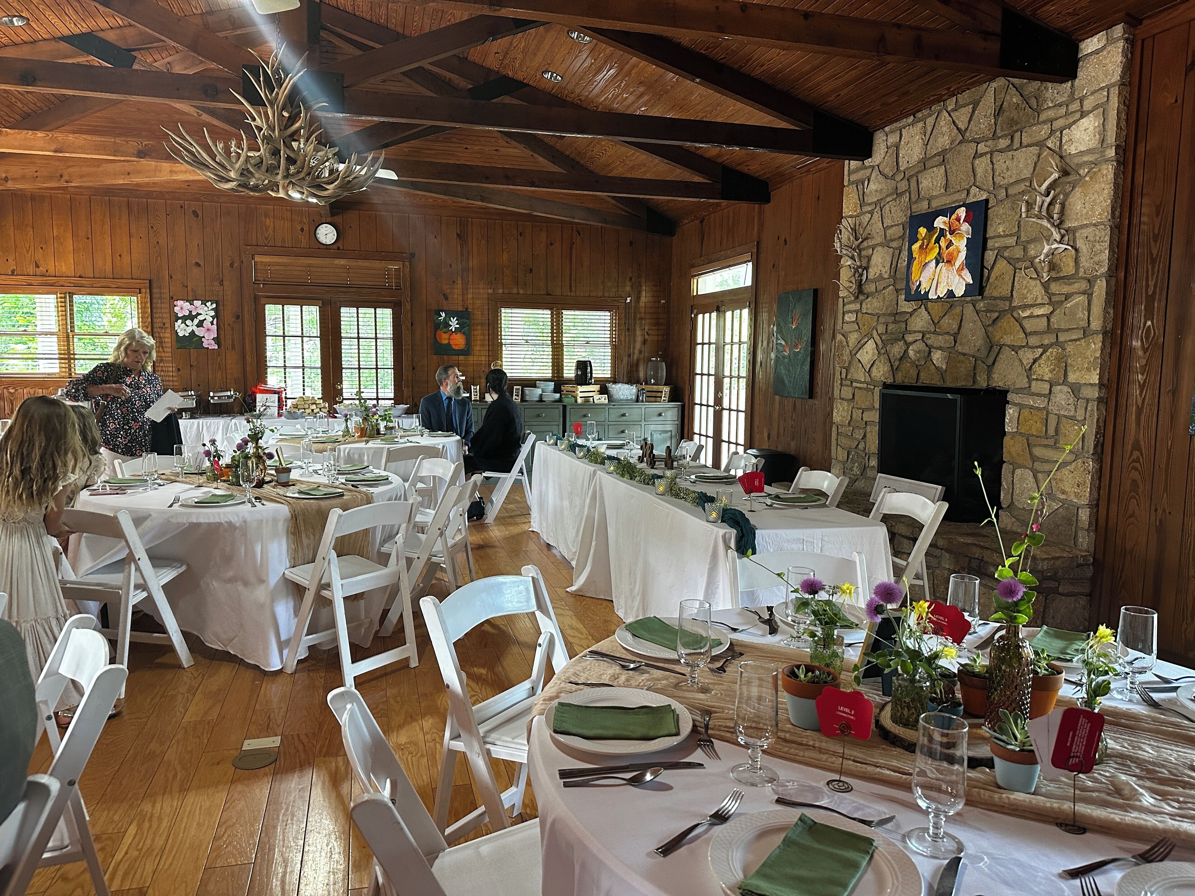 A spacious dining area with high ceilings and wooden beams, featuring stone walls and a fireplace. Tables are set with white tablecloths and green napkins, and there are potted plants on the tables.