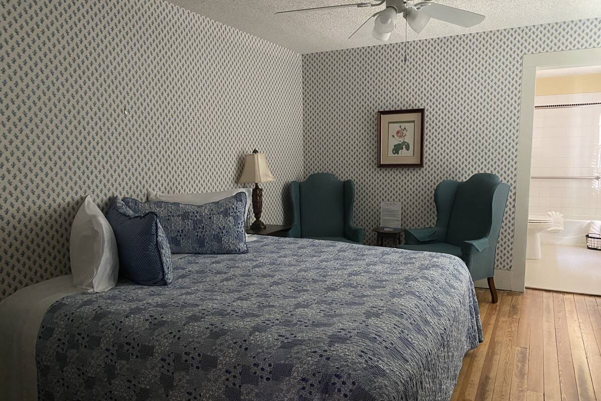 A bedroom with a patterned bedspread, two green chairs, a ceiling fan, and a framed picture on the wall.