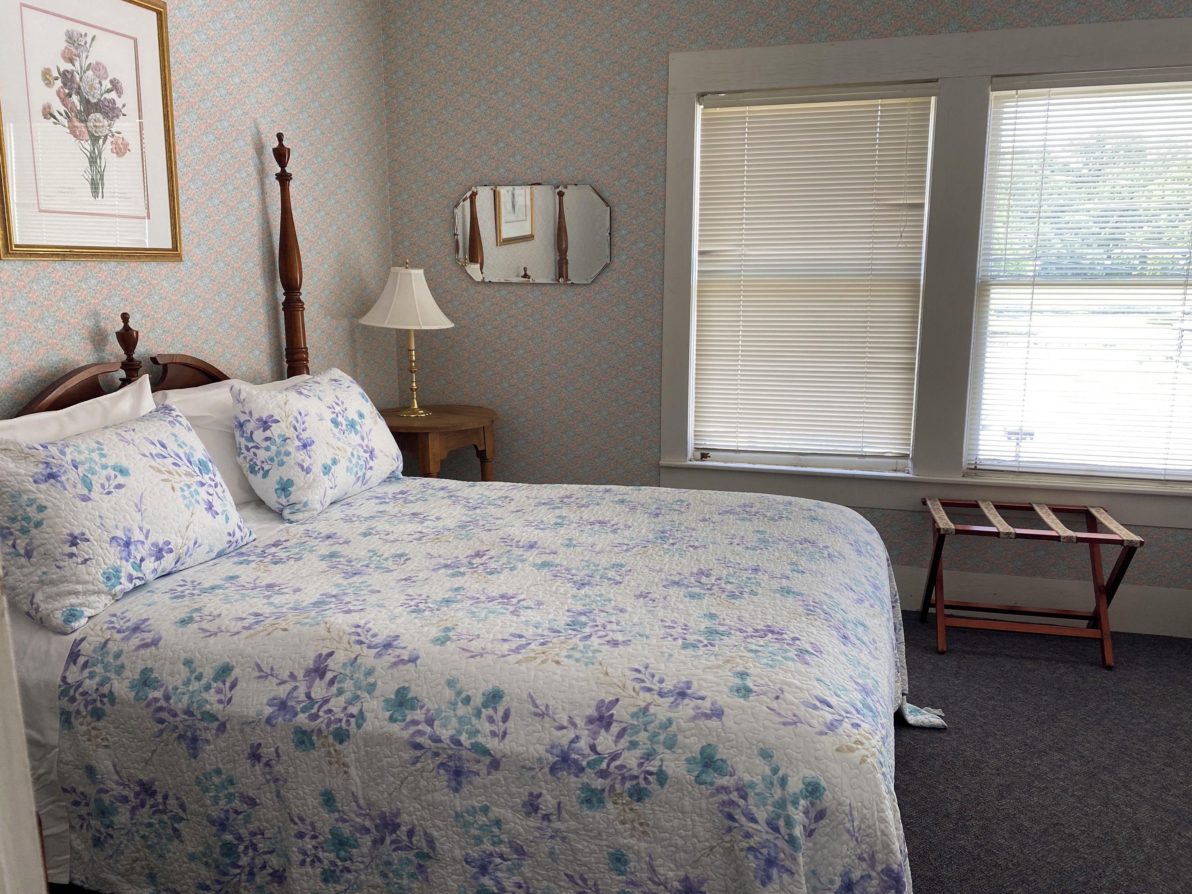 Une chambre à coucher avec une couvre-lit à motifs floraux, une tête de lit en bois, une table de chevet avec une lampe, un miroir et une fenêtre avec des stores.