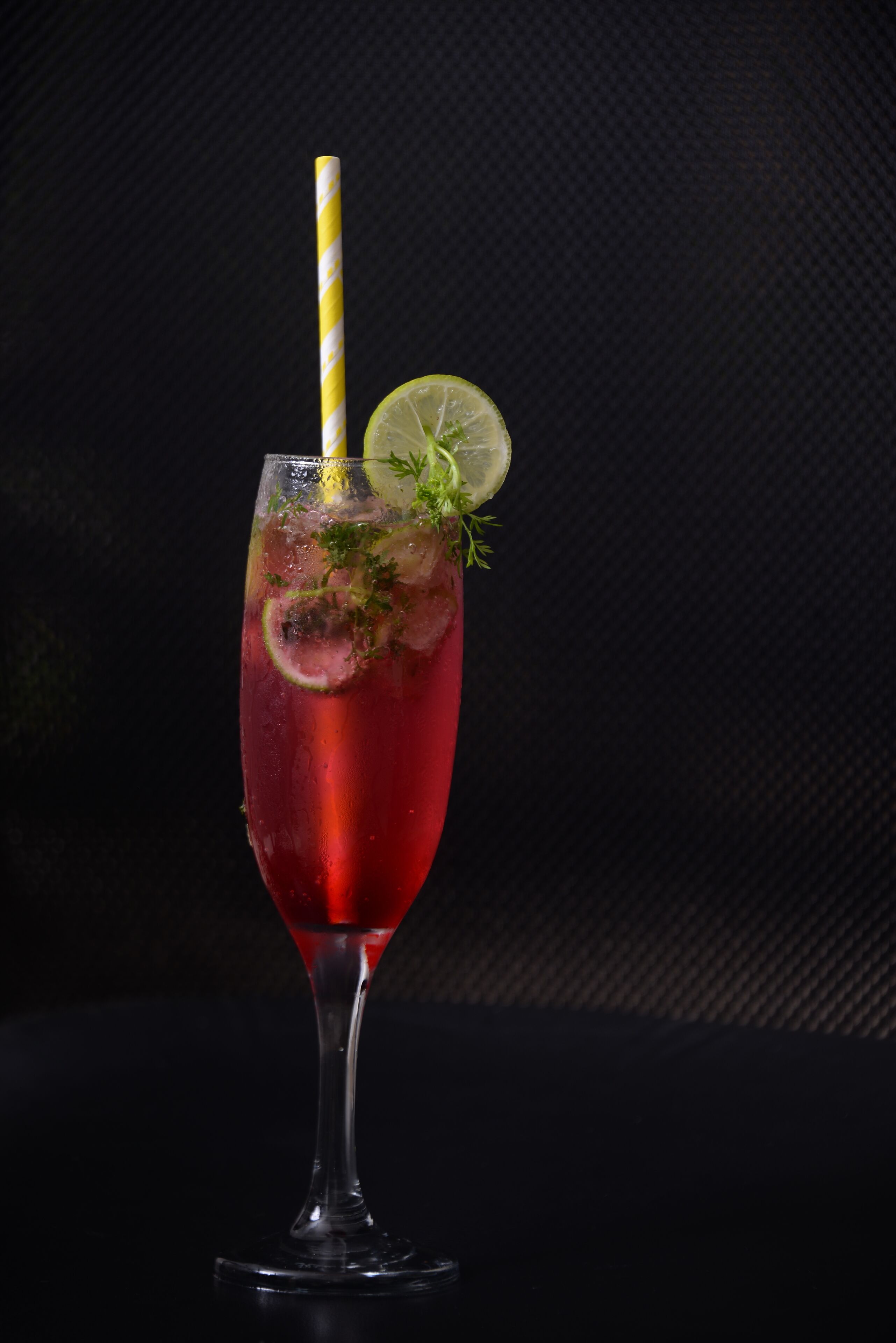 A cocktail glass with a yellow and white striped straw, garnished with a slice of lime and a sprig of herbs, set against a dark background.