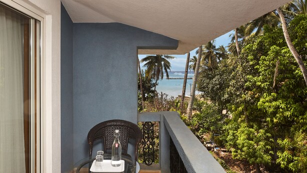 A balcony with a view of the ocean and palm trees, a wicker chair, and a small table with a bottle and glasses. - The Hawaii Comforts (Panaji)
