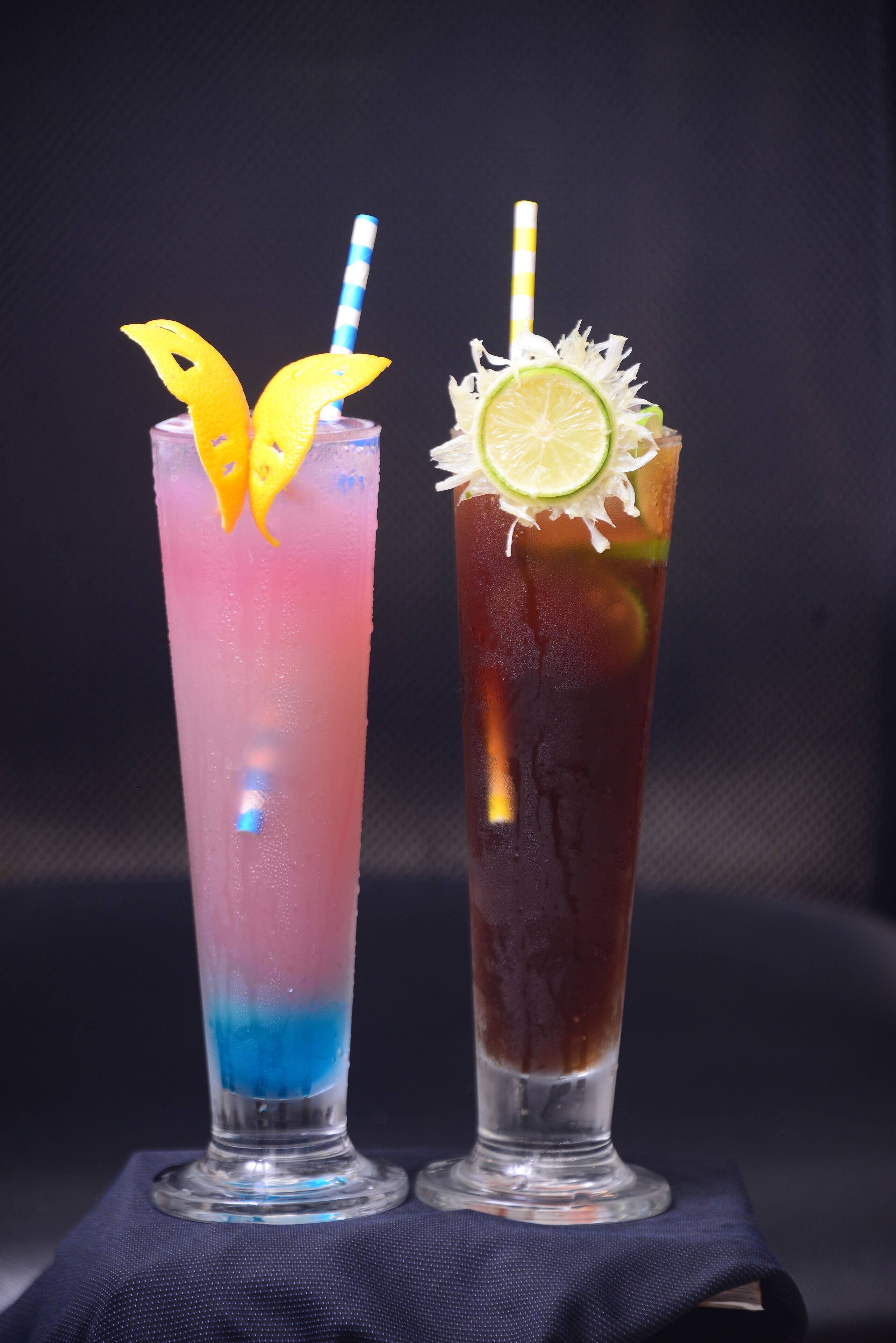 Dos vasos altos de cócteles con pajitas, uno con un degradado azul y rosa y el otro de color oscuro y adorno de lima.