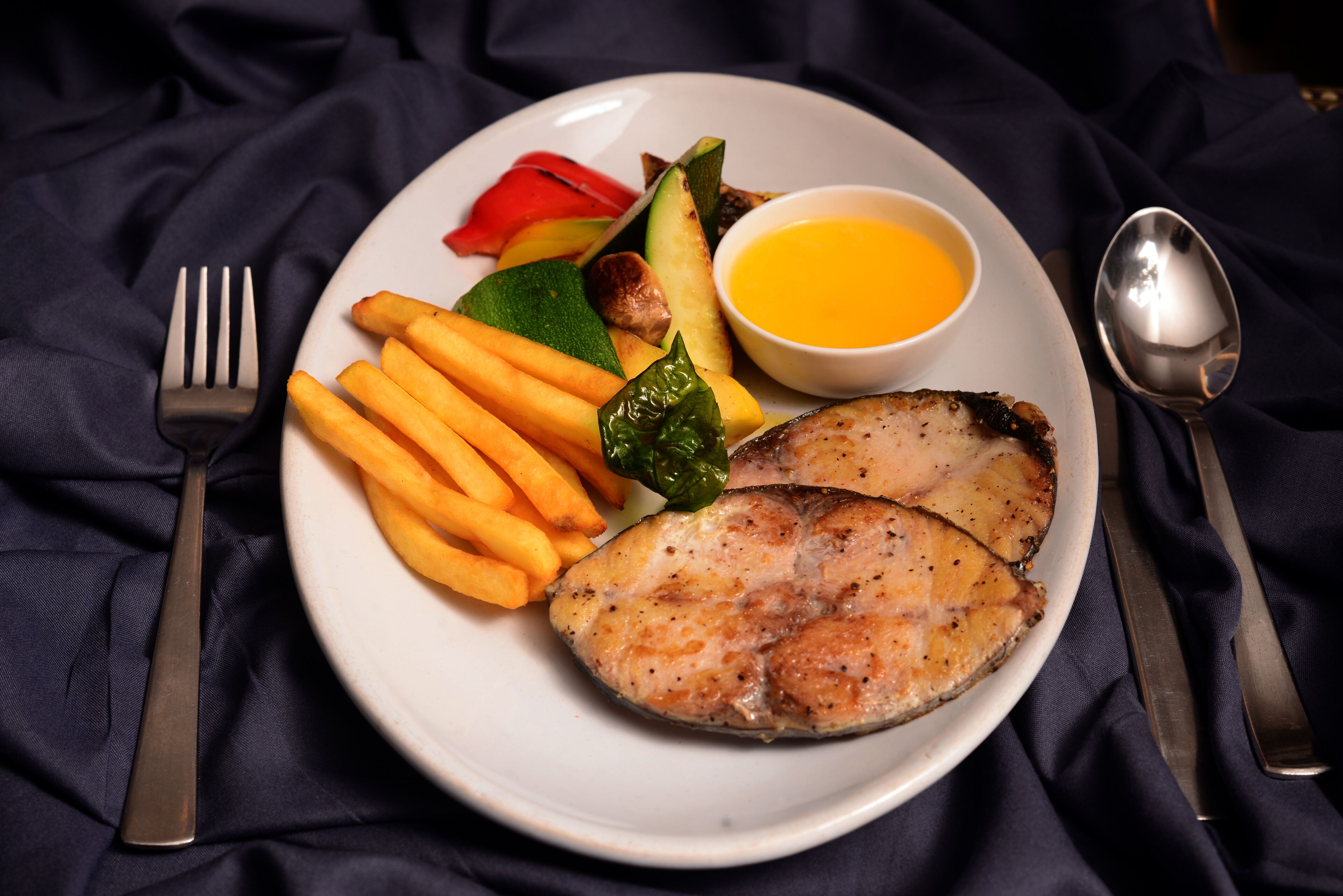 A plate with grilled fish, French fries, vegetables, and a small bowl of sauce.
