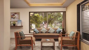 A modern living room with a brown sofa, two wooden armchairs, a coffee table, and a large window with a view of greenery. - The Hawaii Comforts (Panaji)