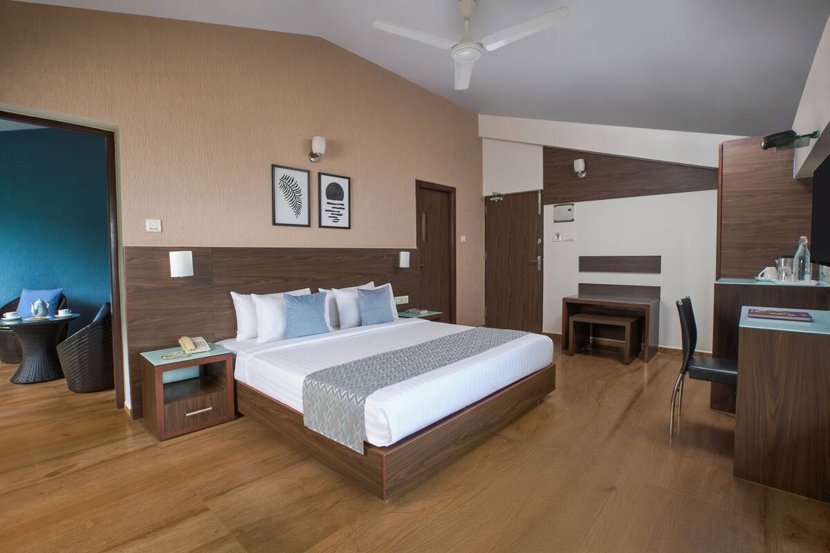 A hotel room with a large bed, a desk, a chair, a small table with a phone, and a wall-mounted light.