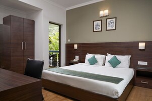Habitación de hotel con una cama grande, un escritorio y un armario. Hay luces fijadas en la pared y una ventana con vistas a la vegetación.