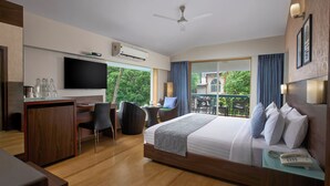 A modern hotel room with a large bed, a desk with a flat-screen TV, a balcony with outdoor seating, and an air conditioning unit. - The Hawaii Comforts (Panaji)