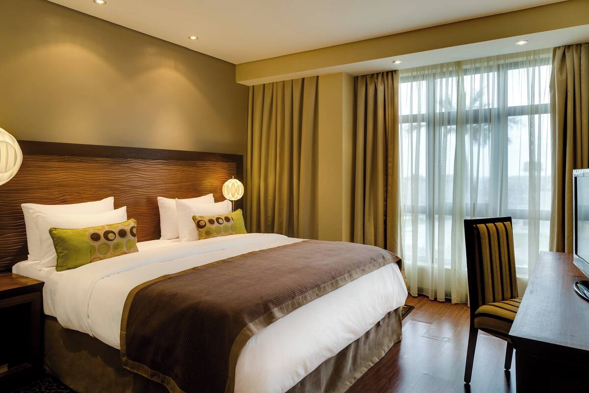 A hotel room with a large bed, a wooden headboard, a desk with a chair, and a large window with curtains.