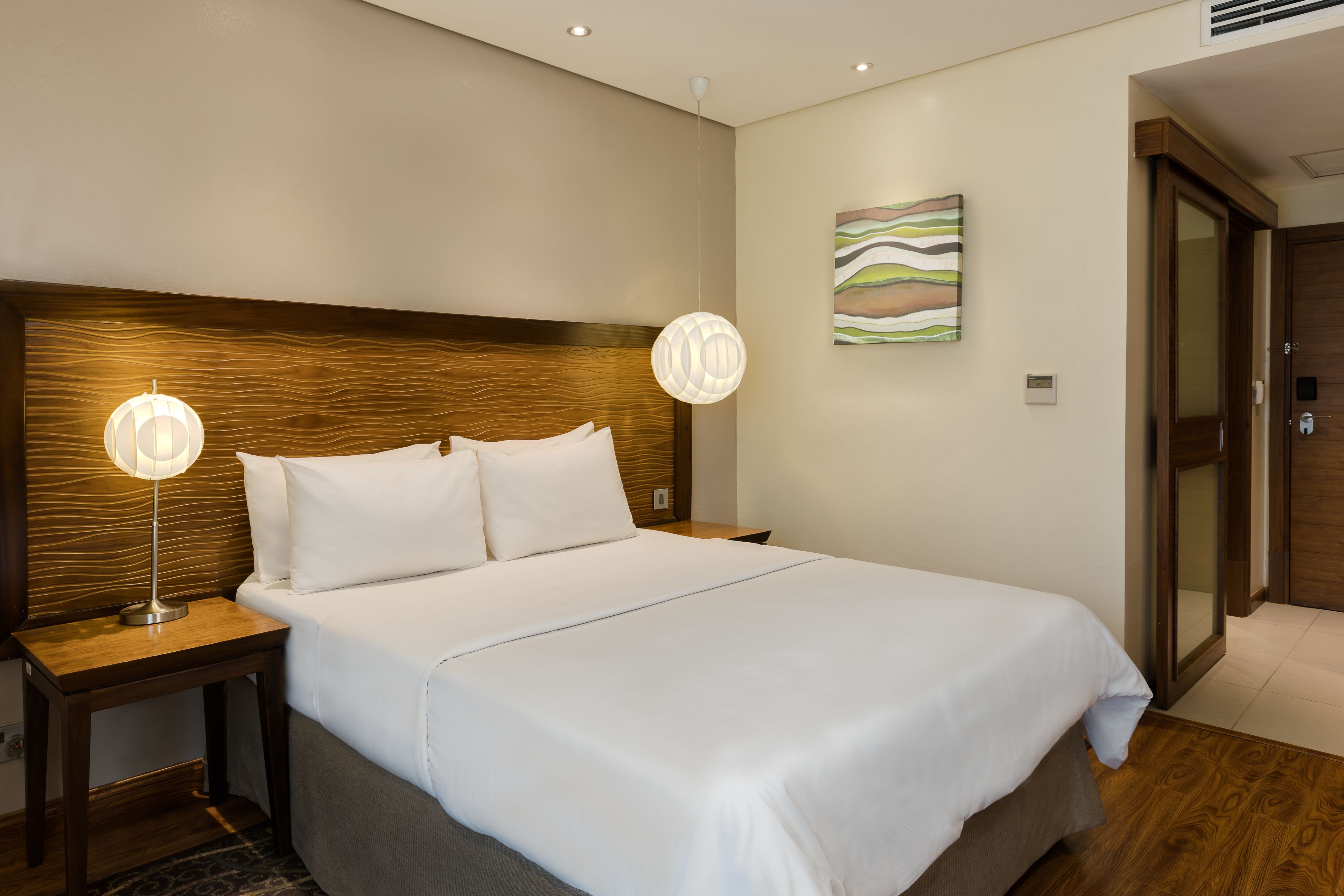 Une chambre d’hôtel avec un grand lit, une tête de lit en bois, deux lampes de chevet et une peinture abstraite moderne accrochée au mur.