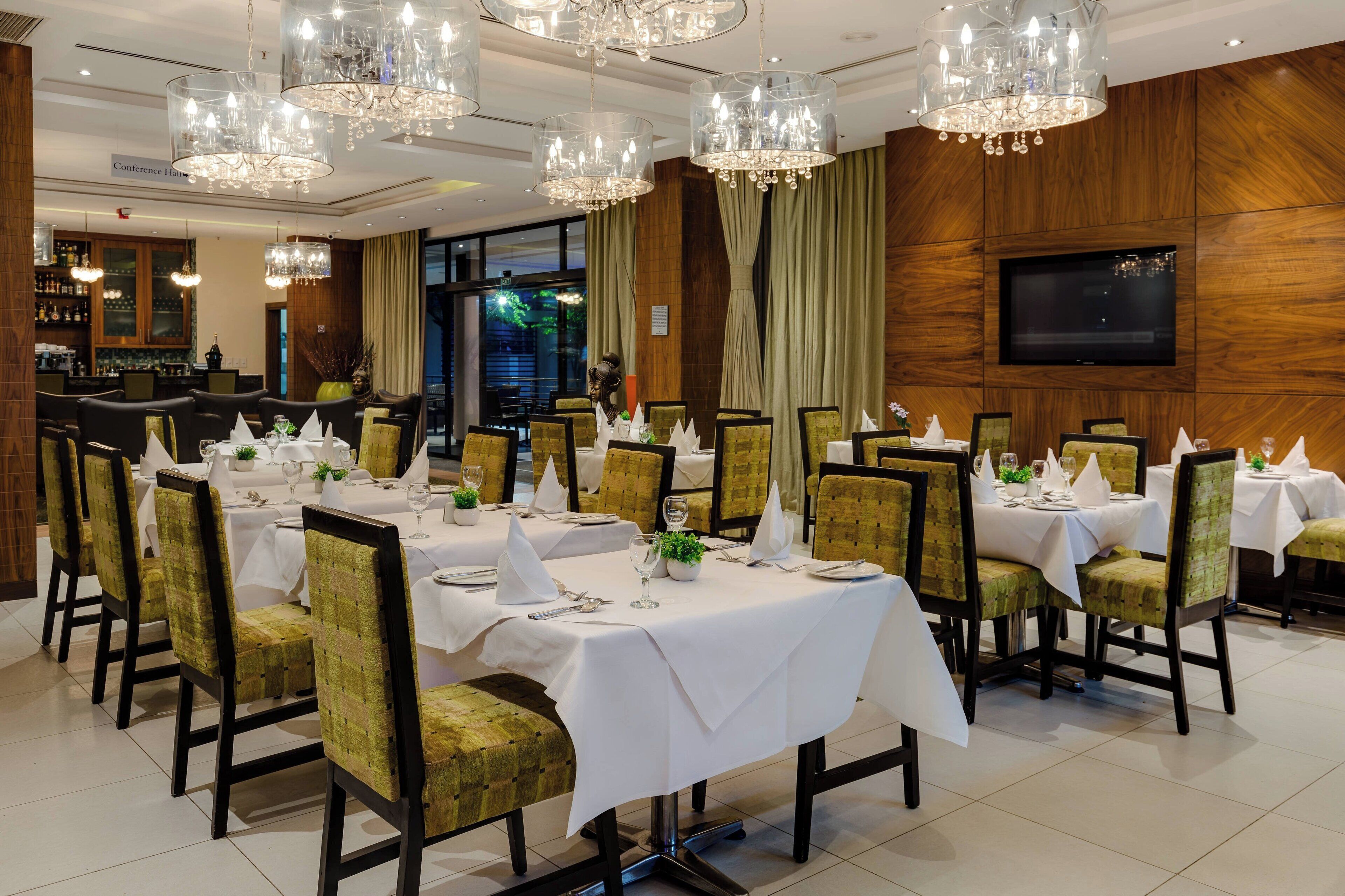 A formal dining room with multiple tables set for service, elegant chandeliers, and a bar area in the background.