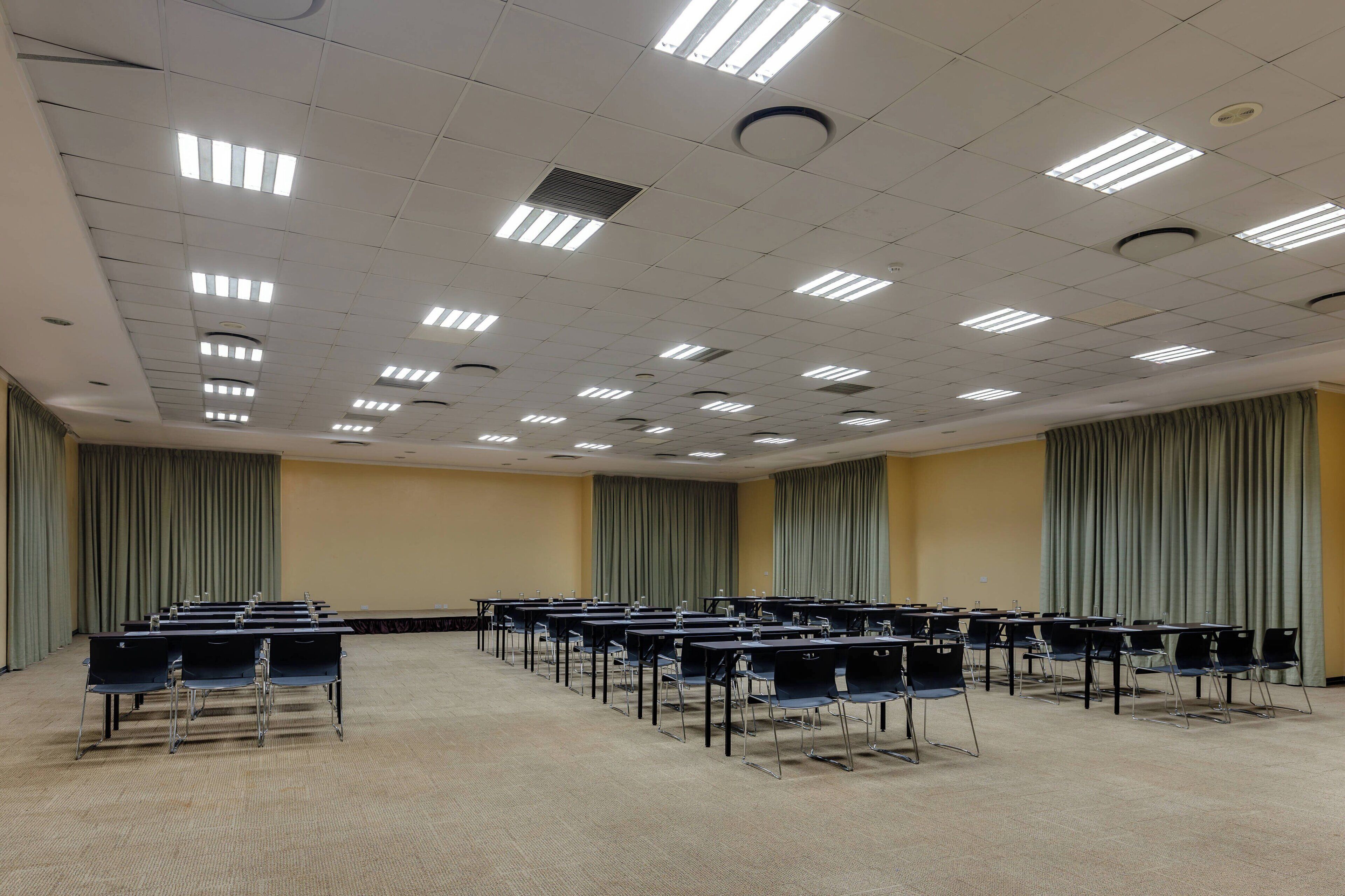 A conference room with rows of tables and chairs, ceiling lights, and curtains.