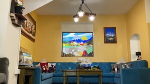 A living room with a blue sofa, wooden flooring, a ceiling fan, and a painting of a mountain landscape. - Kituandes Hotel (Quito)