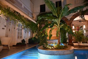 Una piscina interior con borde de azulejos, rodeada de abundante vegetación y un muro de ladrillo.