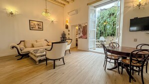 Una sala de estar con sofá, sillas, una mesa de centro, una mesa de comedor con sillas, una araña, televisor y un ventanal con vistas a la vegetación.